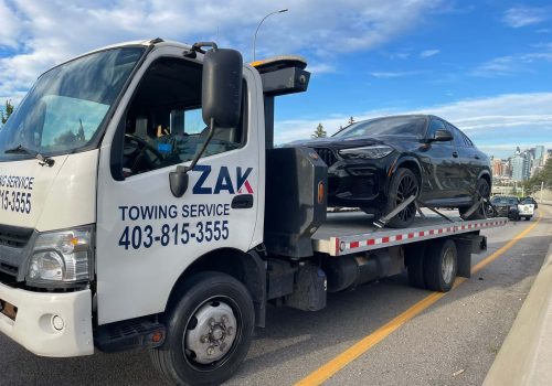 Zak Towing flatbed tow truck transporting a BMW SUV on a Calgary roadway with the city skyline in the background.