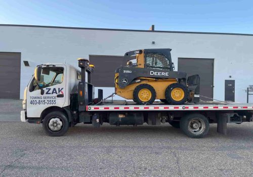 Zak Towing Service is transporting heavy construction equipment on a flatbed tow truck in Calgary, Alberta. The image shows a John Deere skid steer securely loaded and chained for safe commercial equi...Sorry but this string is too long !!!