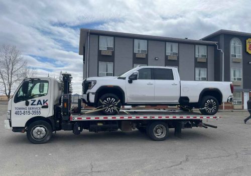 Flatbed tow truck from Zak Towing Service carrying a white pickup truck in front of a Calgary building
