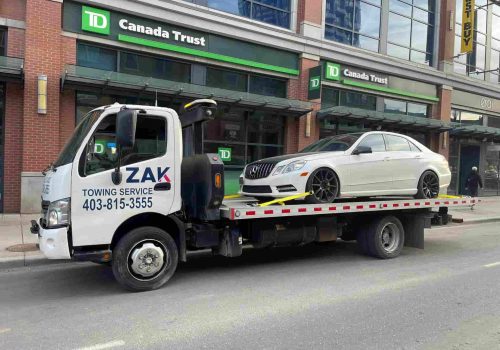 Zak Towing flatbed truck transporting a white luxury car in front of TD Canada Trust in downtown Calgary.